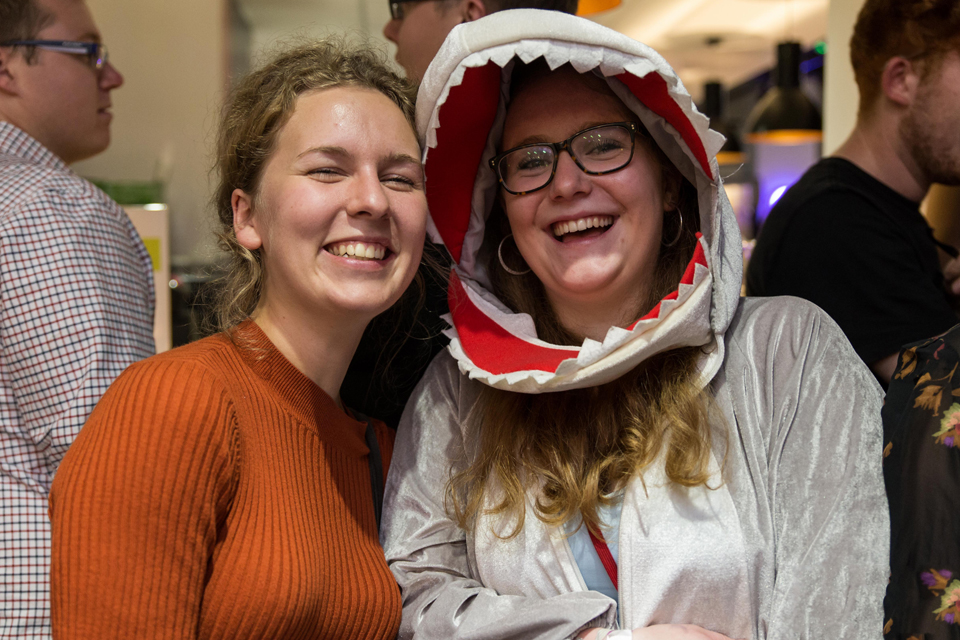 Two female students, one with glasses and in a shark costume, smiling at the camera, with other students chatting behind them.